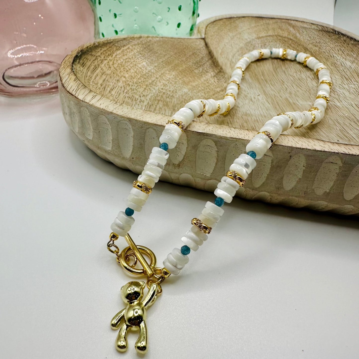A mother of pearl, gold, and teal beaded necklace with a gold toggle clasp and a gold balloon teddy bear charm lies draped over a wooden dish on a white table. There are two colored glass vases in the background.
