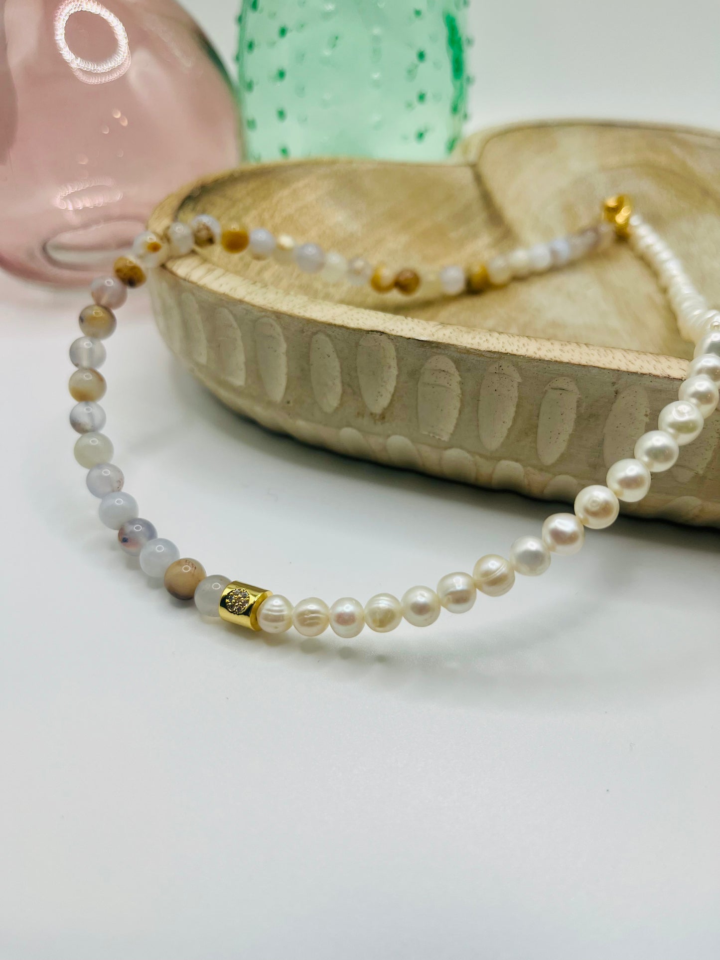 A necklace of half pearls, half blue chalcedony lies draped over a wooden dish on a white table. There are two colored glass vases in the background.