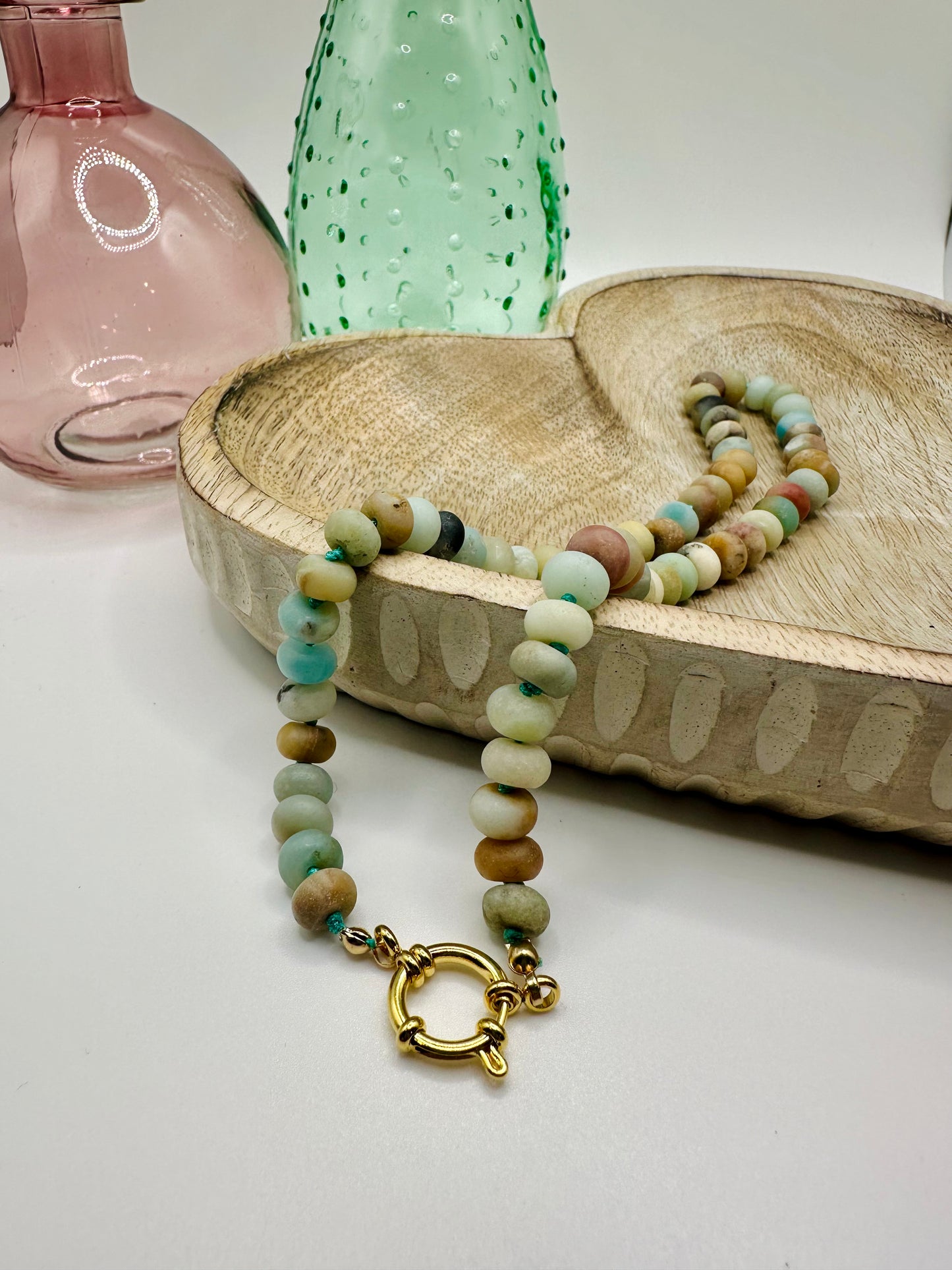 A necklace of hand-knotted large amazonite matte stones lies draped over a wooden dish on a white table. There are two colored glass vases in the background.