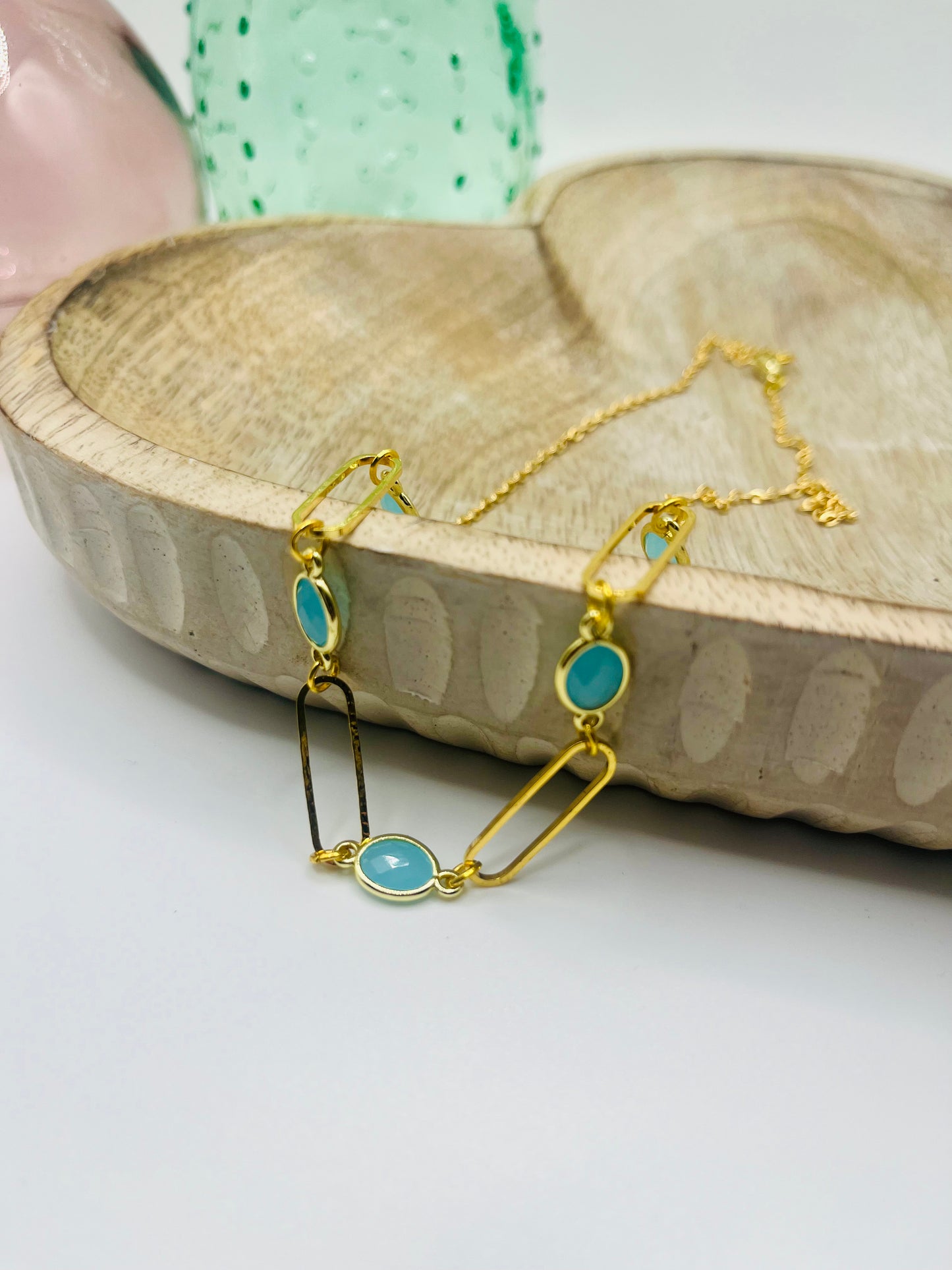 A gold paperclip chain and Austrian crystal necklace lies draped over a wooden dish on a white table. There are two colored glass vases in the background.