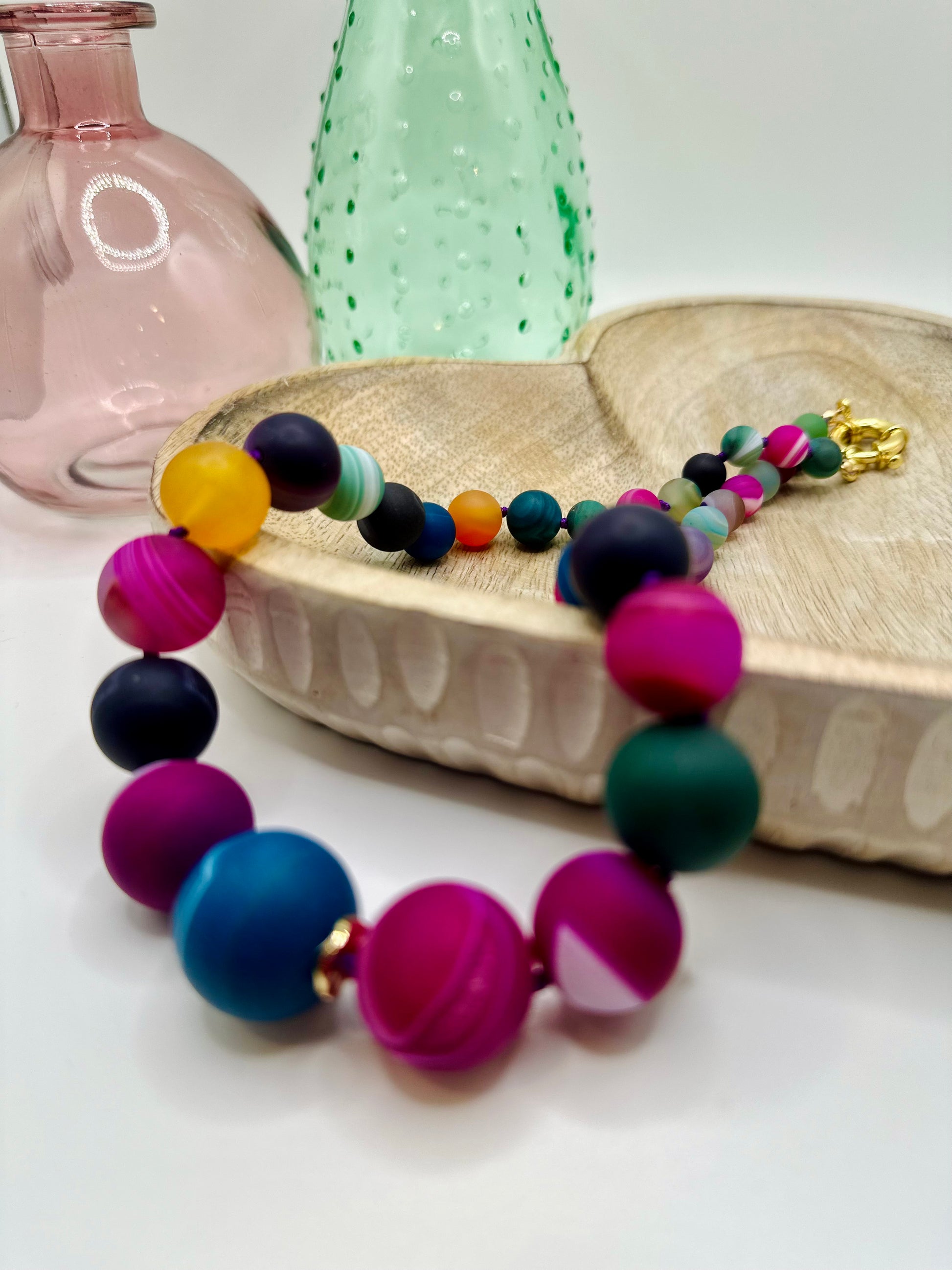 A large chunky stone rainbow agate necklace lies draped over a wooden dish on a white table. There are two colored glass vases in the background.