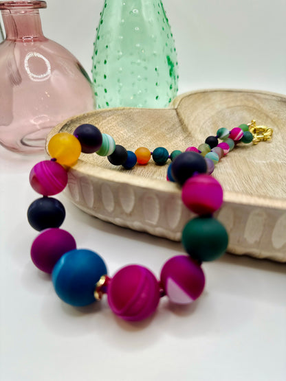 A large chunky stone rainbow agate necklace lies draped over a wooden dish on a white table. There are two colored glass vases in the background.