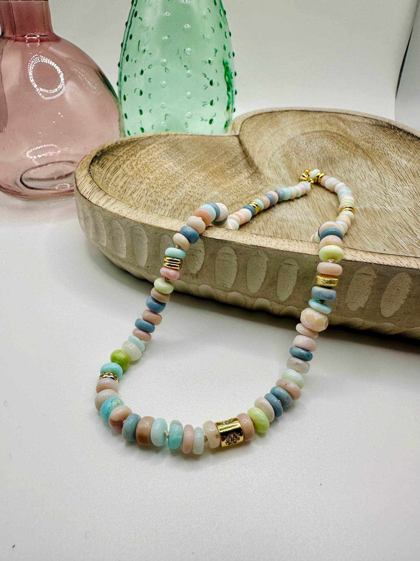 A necklace of hand-knotted Ethiopian opals in pastel shades lies draped over a wooden dish on a white table. There are two colored glass vases in the background.
