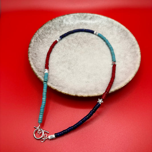 A necklace of dyed turquoise beads in shades of teal, navy, red and white with two sterling silver star accent beads lies in a clay plate with a bright red background