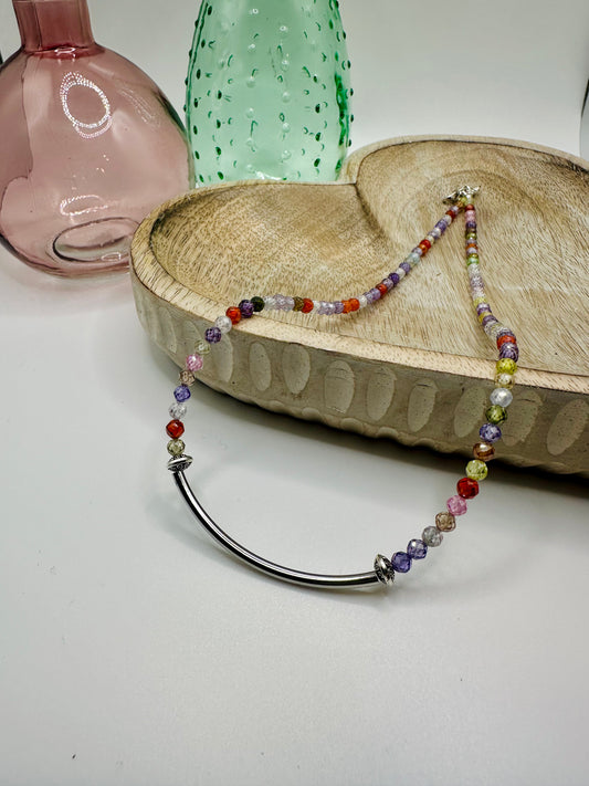 A necklace with multi-colored cubic zirconia crystals and a silver rounded bar lies draped over a wooden dish on a white table. There are two colored glass vases in the background.