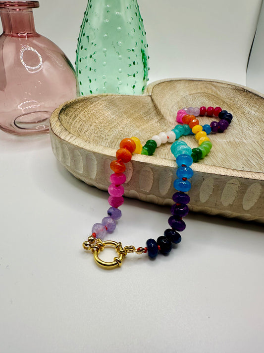 A rainbow of candy jade is hand-knotted on this necklace which lies draped over a wooden dish on a white table. There are two colored glass vases in the background.