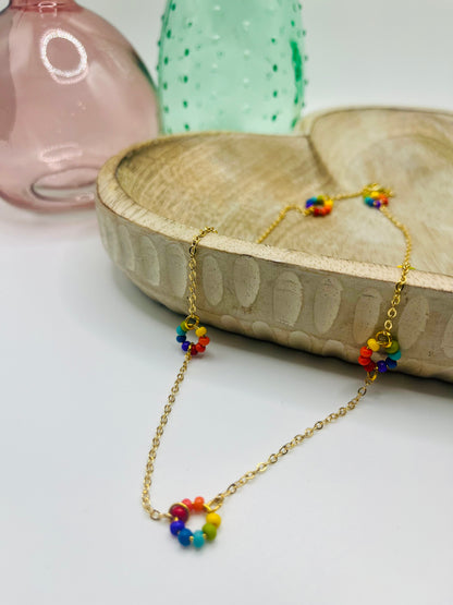 A gold chain and small round rainbow seed bead necklace lies draped over a wooden dish on a white table. There are two colored glass vases in the background.