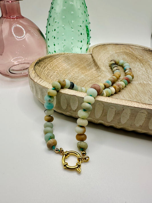 A necklace of hand-knotted large amazonite matte stones lies draped over a wooden dish on a white table. There are two colored glass vases in the background.