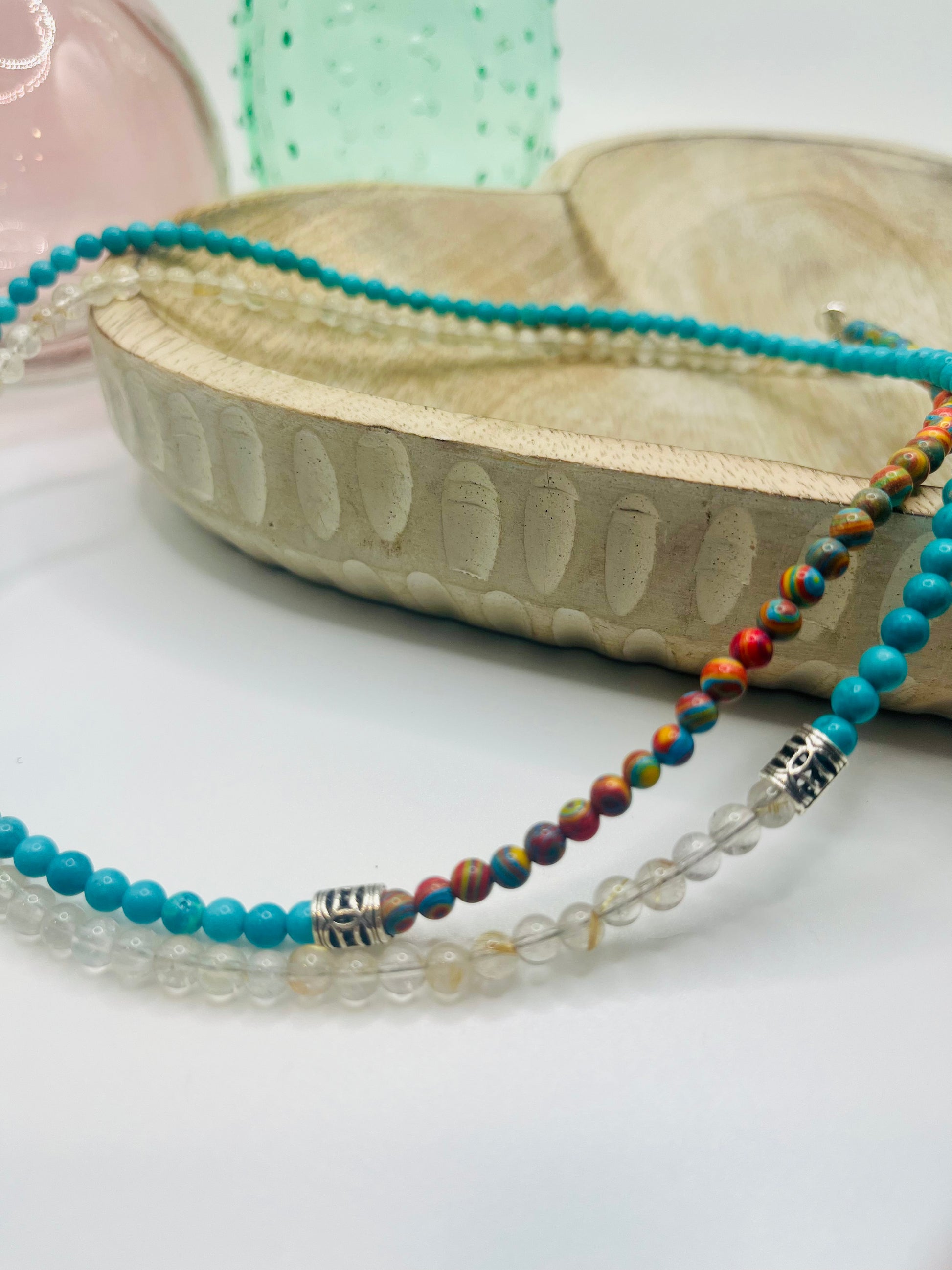 A double-wrapped beaded necklace in rutilated quartz, turquoise and other colorful beads lies draped over a wooden dish on a white table. There are two colored glass vases in the background.