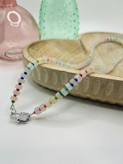 A clear quartz and rainbow seed bead necklace lies draped over a wooden dish on a white table. There are two colored glass vases in the background.