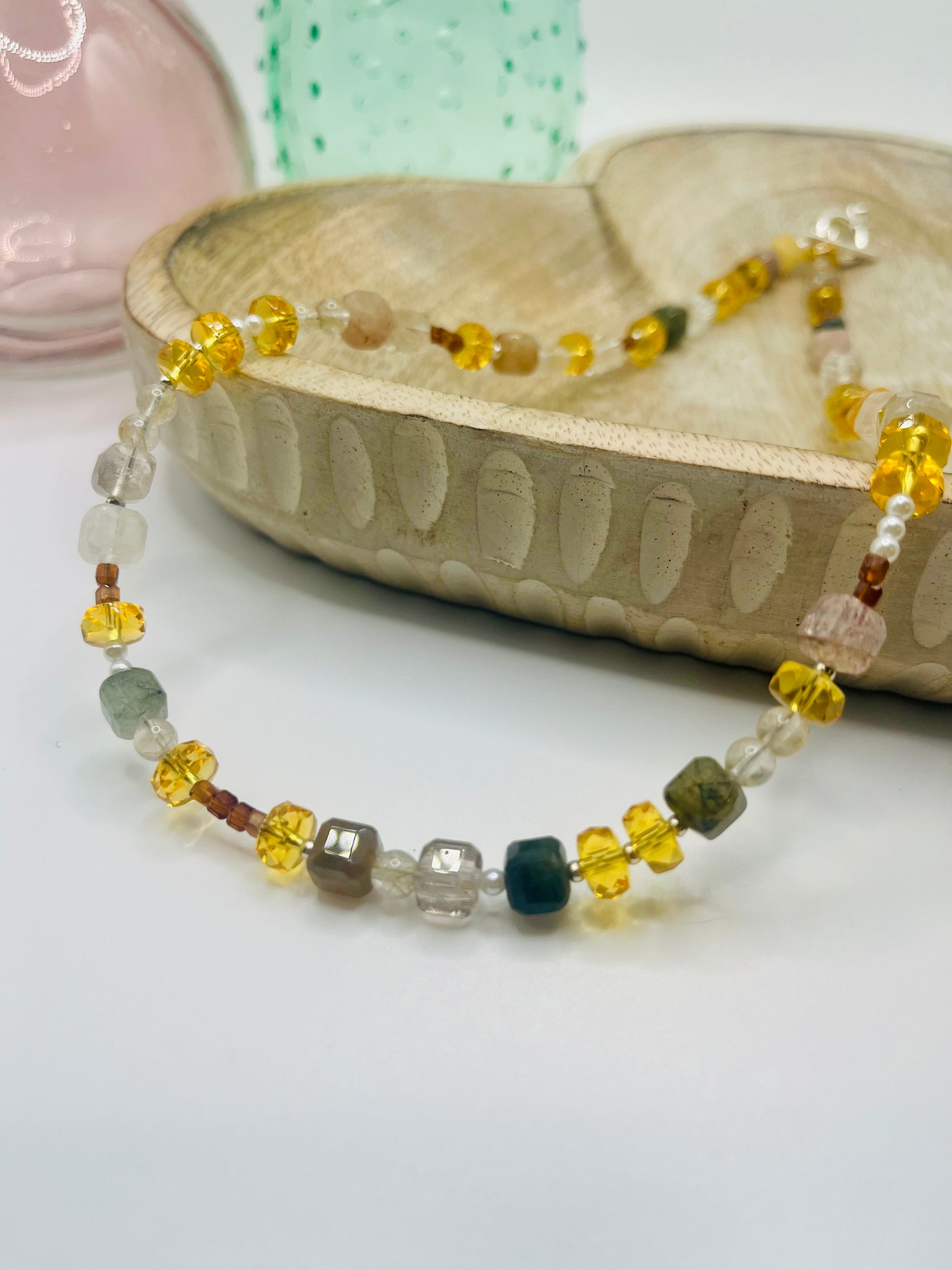 A necklace strung of citrine, quartz, pearls, hessonite garnet and more lies draped over a wooden dish on a white table. There are two colored glass vases in the background.