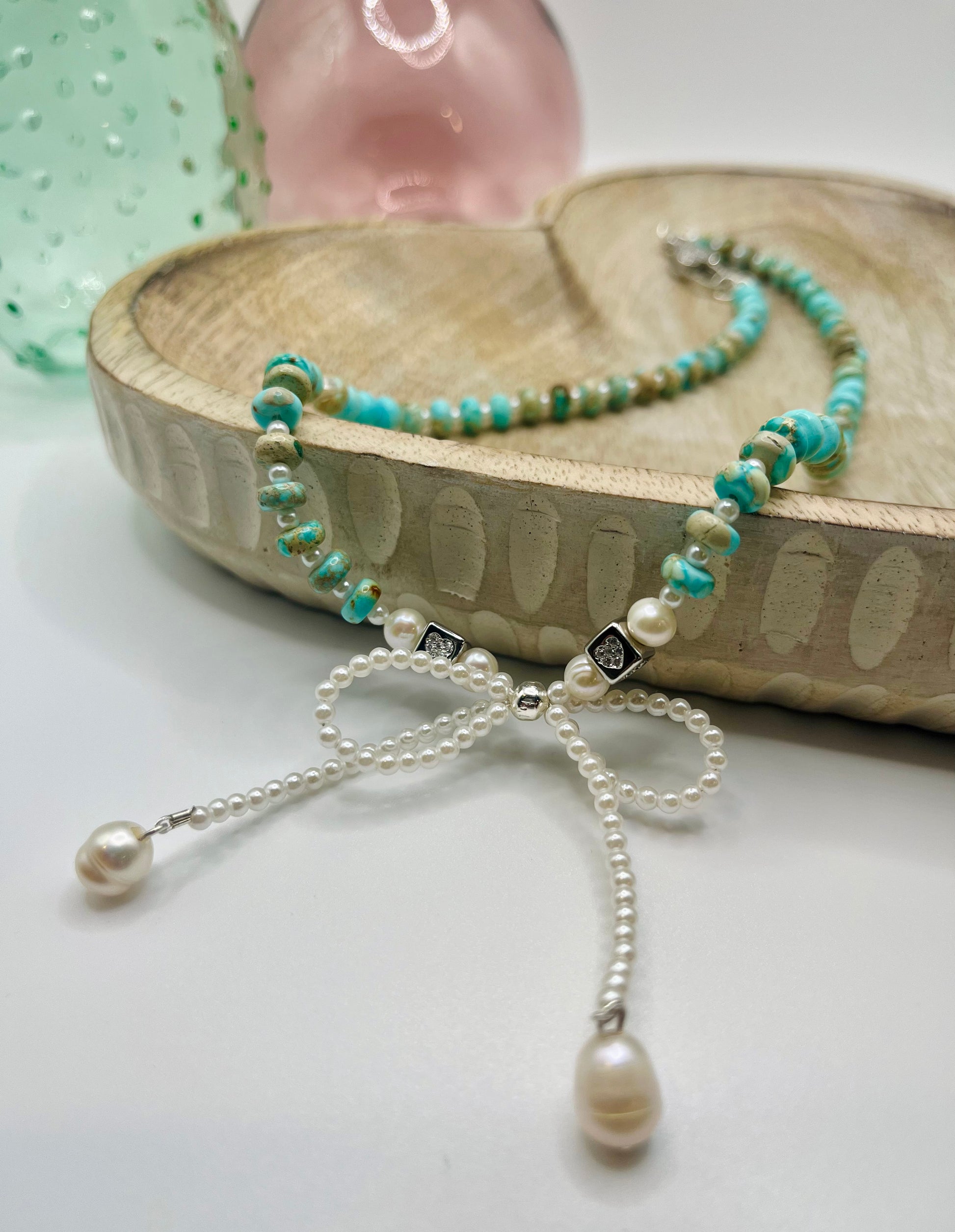 A necklace of turquoise and pearls with a prominent pearl bow lies draped over a wooden dish on a white table. There are two colored glass vases in the background.