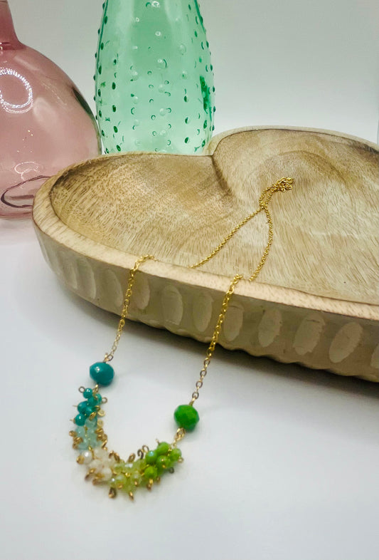 A chain necklace with teal, white, lime and green cubic zirconia crystals lies draped over a wooden dish on a white table. There are two colored glass vases in the background.