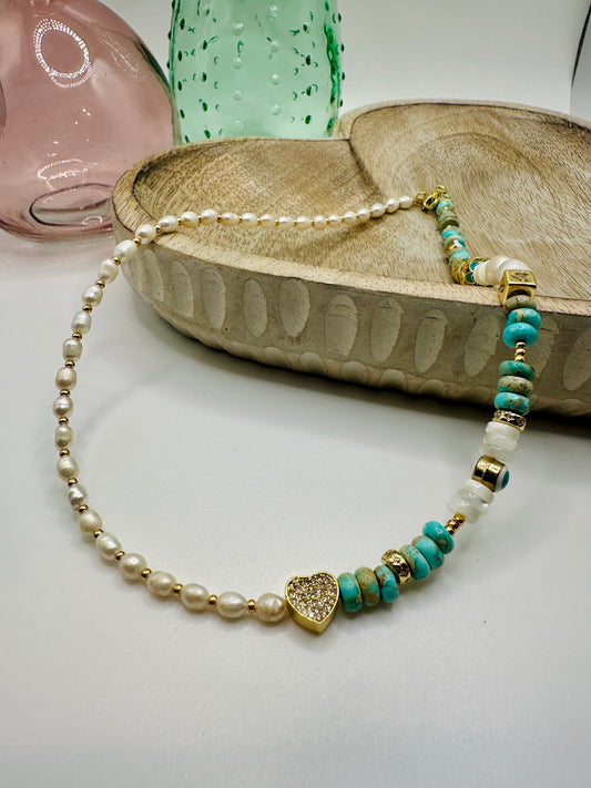 A necklace of half seed pearls and half mixture of mother of pearl, turquoise, and gold findings lies draped over a wooden dish on a white table. There are two colored glass vases in the background.