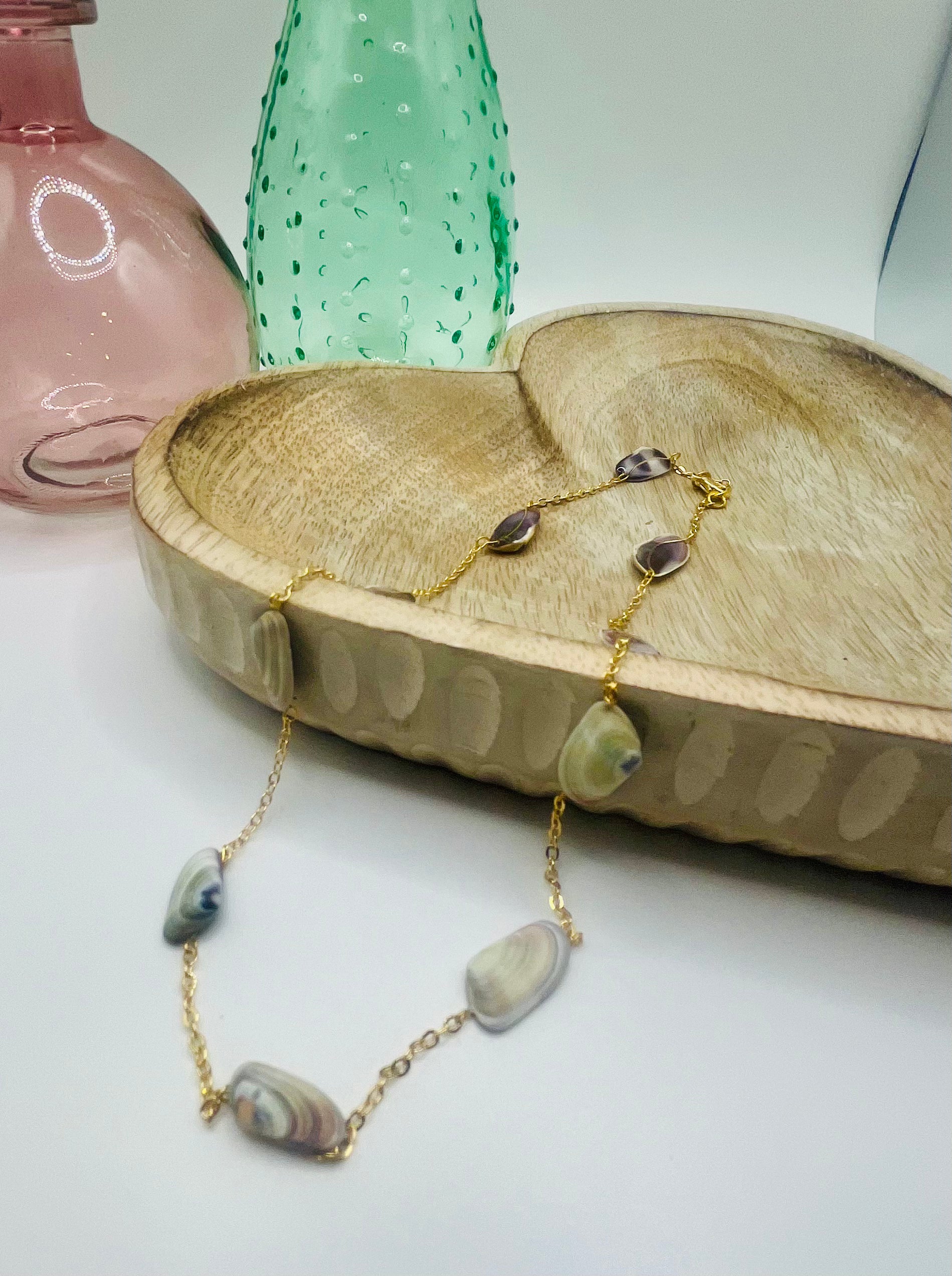 A gold chain and purple small shell necklace lies draped over a wooden dish on a white table. There are two colored glass vases in the background.